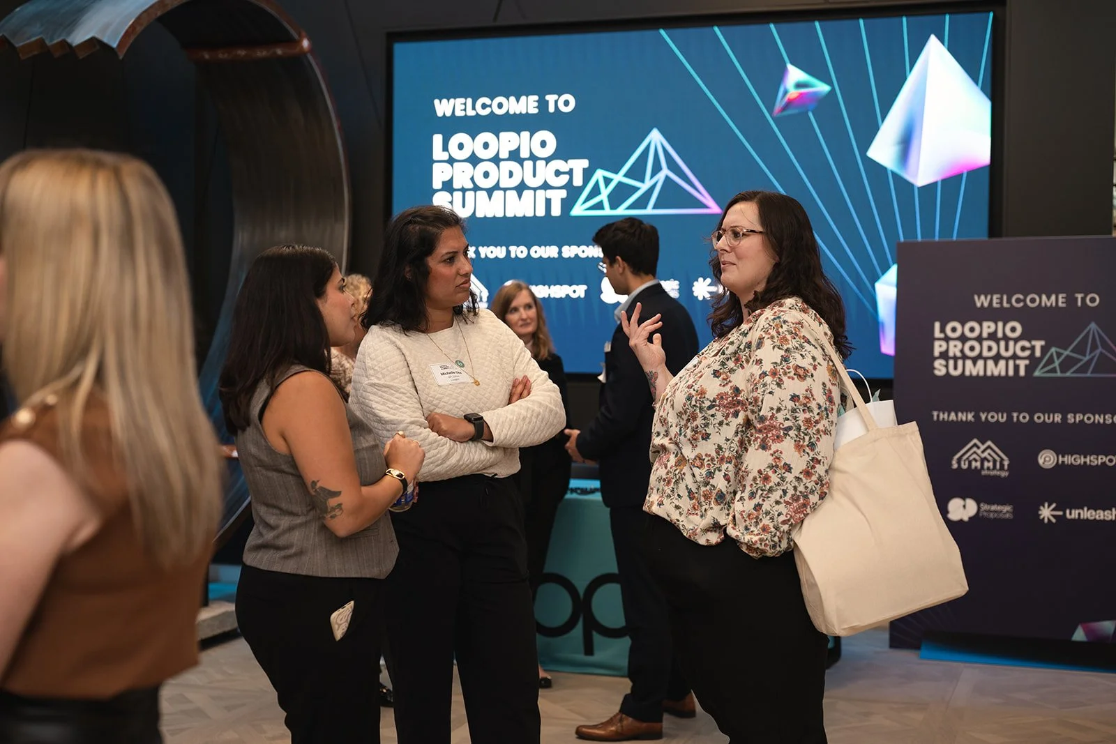 Group of people engaged in conversation at the Loopio Product Summit with a digital screen in the background displaying event details.