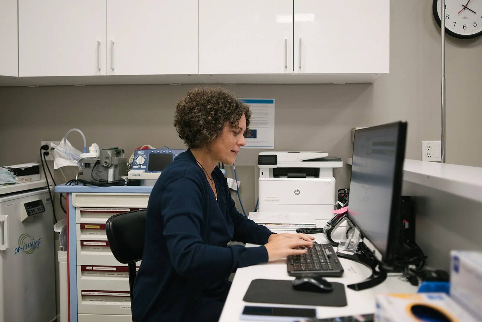 A healthcare worker sitting at a desk in a medical office, working on a computer with medical equipment and supplies around her.