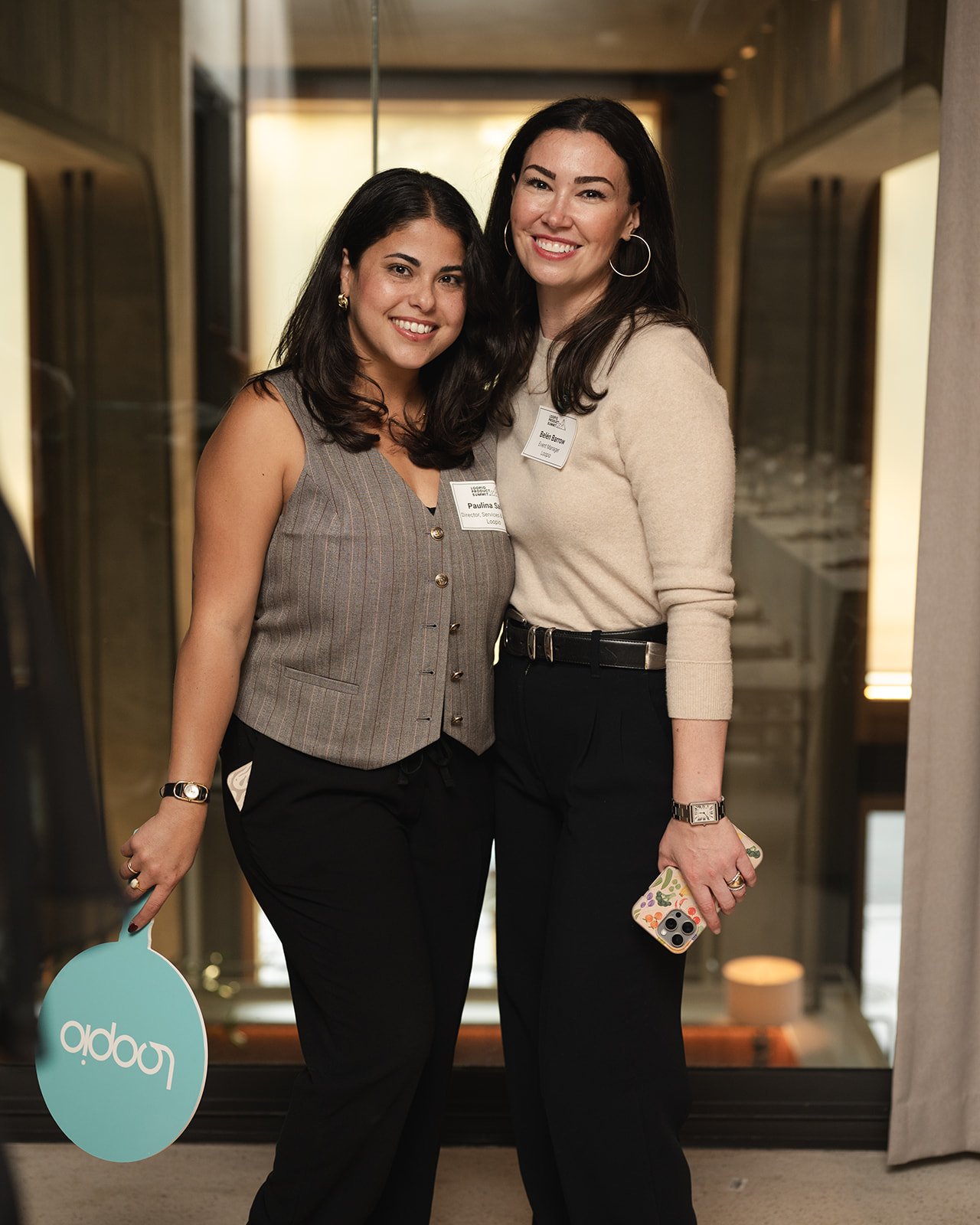 Two women smiling and standing close together in an indoor setting, both wearing business casual attire and name tags.