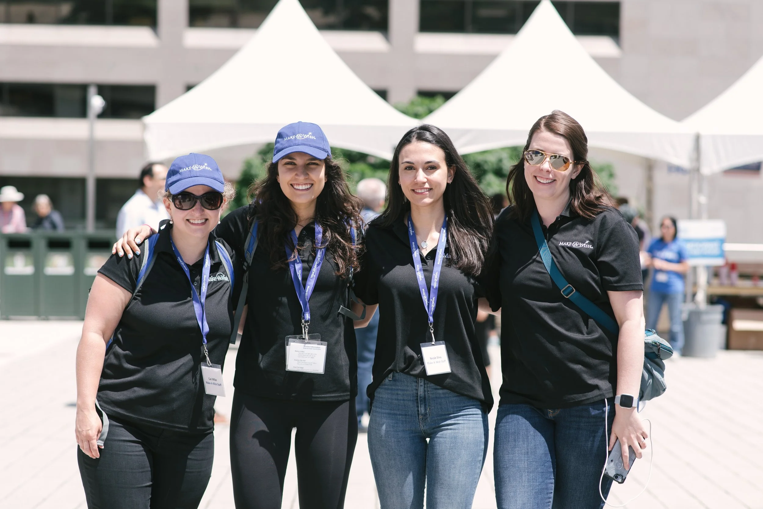 Four women wearing Make-A-Wish Foundation shirts and lanyards standing outdoors at a sunny event, smiling for the camera.