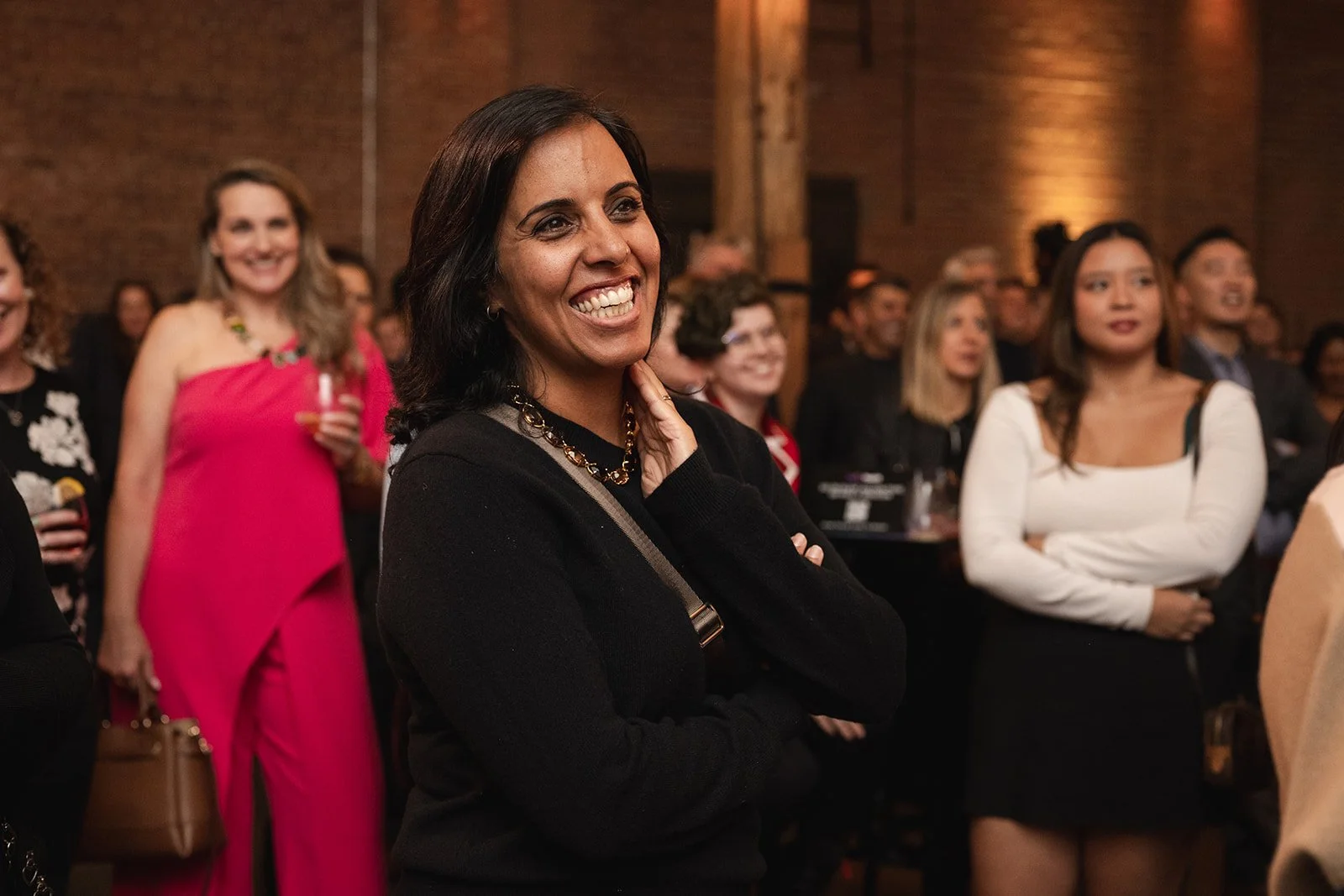 A group of women at a corporate event, with a woman in the foreground smiling and touching her face, dressed in a black top with a gold necklace. The background shows other women and a brick wall.