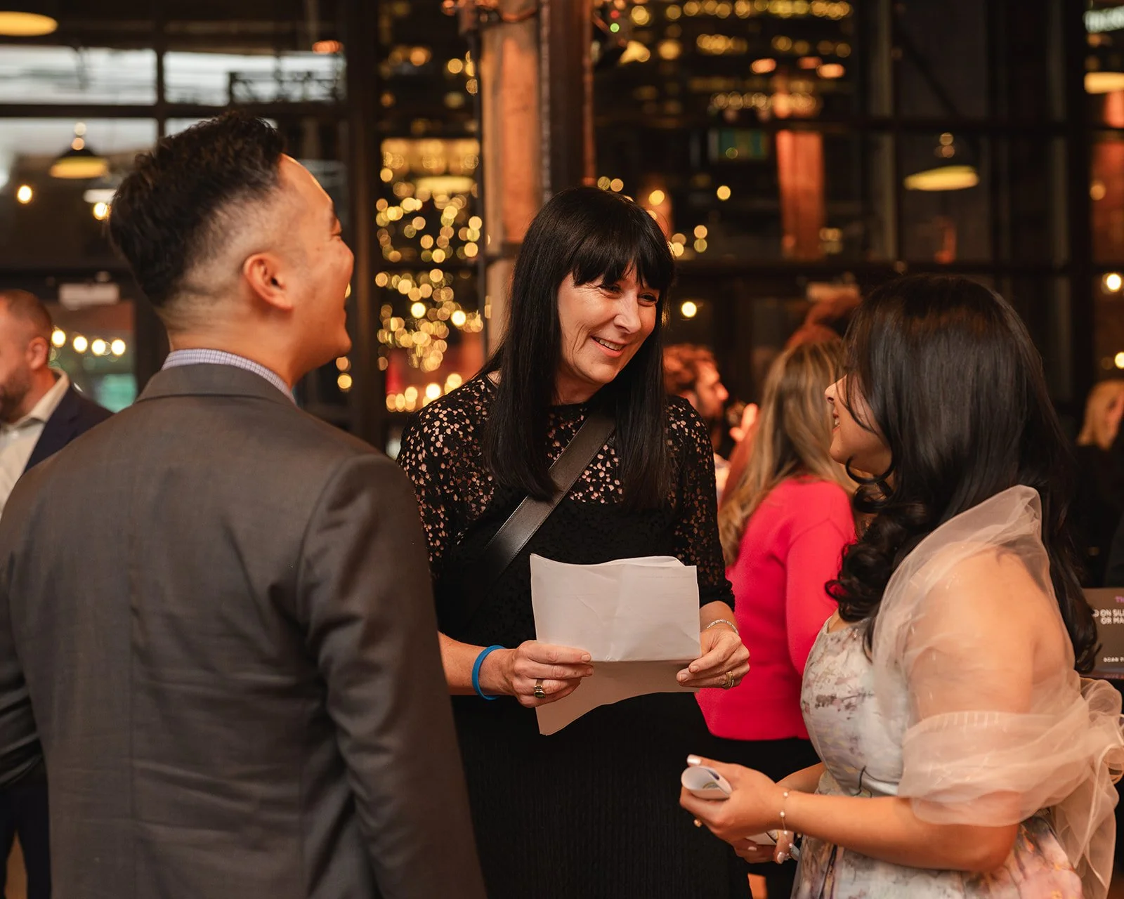 Three people, two women and one man, are talking and smiling at an indoor social gathering with warm lighting and decorative string lights in the background.