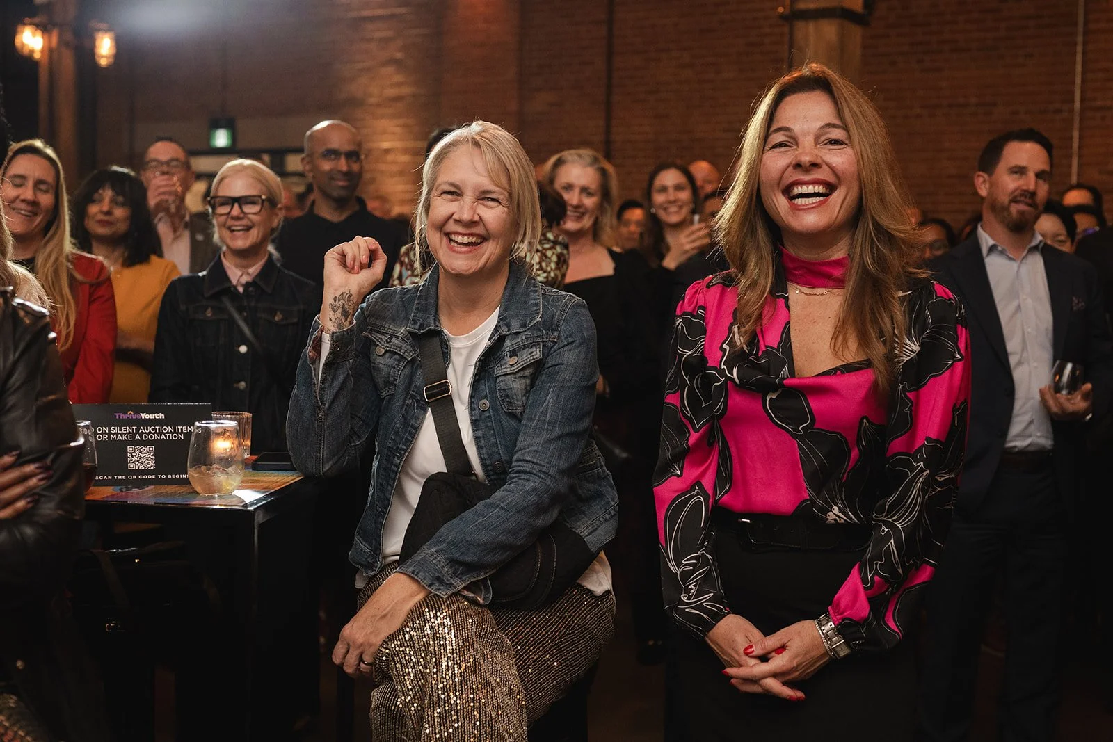 A group of people at an indoor event, with two women in the front smiling and laughing. One woman wears a denim jacket, white shirt, and sequin pants, while the other wears a pink and black patterned blouse. Others in the background are also smiling 