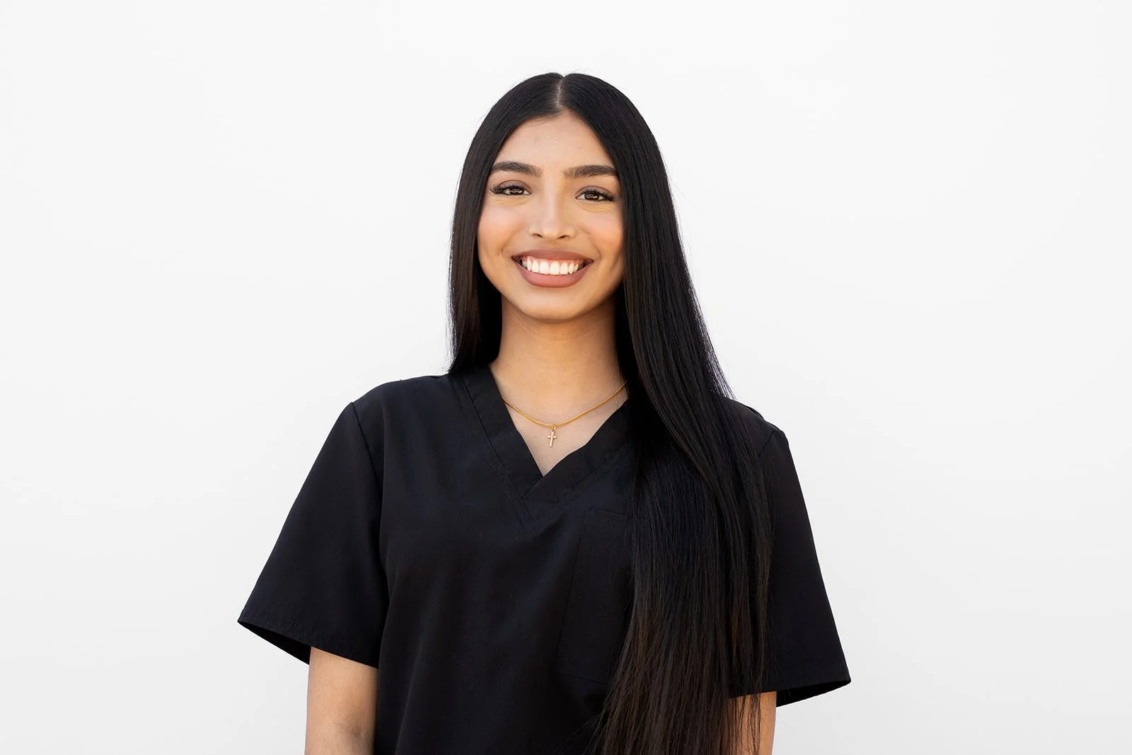 A headshot of a young woman with long black hair, smiling, wearing a black scrubs top and a delicate gold necklace with a cross pendant, standing against a plain white background.