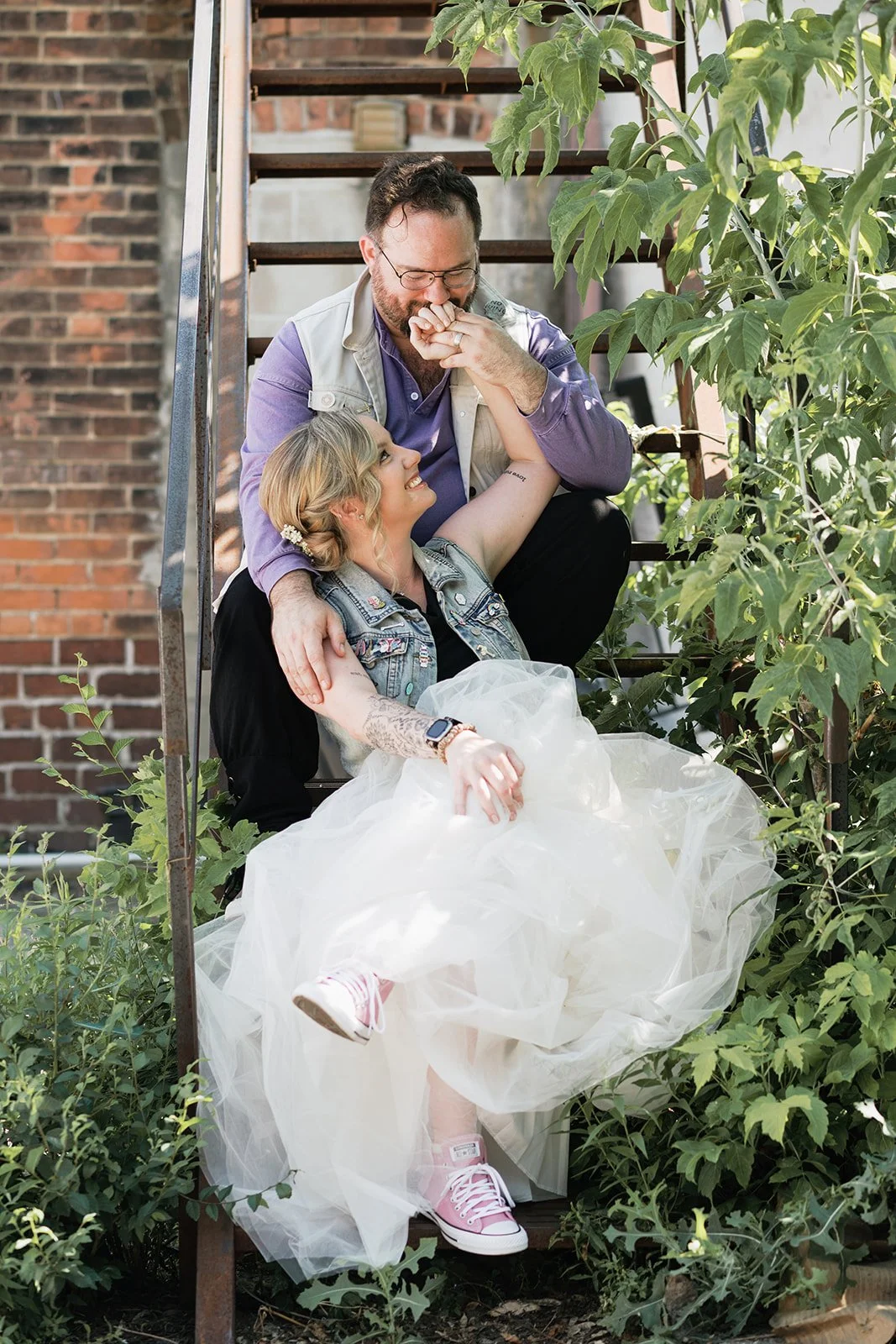 A bride and groom sitting on fire escape stairs, surrounded by green plants, sharing a joyful moment. The woman is wearing a white tu-tu like dress and pink sneakers, while the man is leaning over her, kissing her hand and smiling.