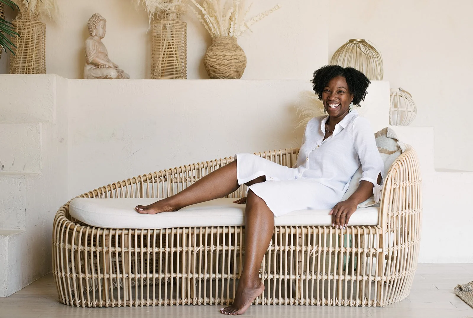 A woman in a white dress sits on a wicker couch in a bright, neutral-toned room, smiling and relaxing at Mintroom Studios in Toronto.