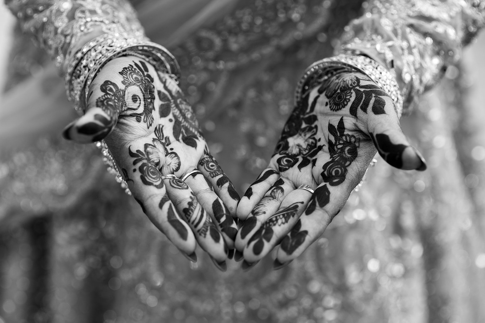 Close-up of two hands with intricate henna designs, adorned with multiple bangles and rings, forming a heart shape against a blurred background.  
