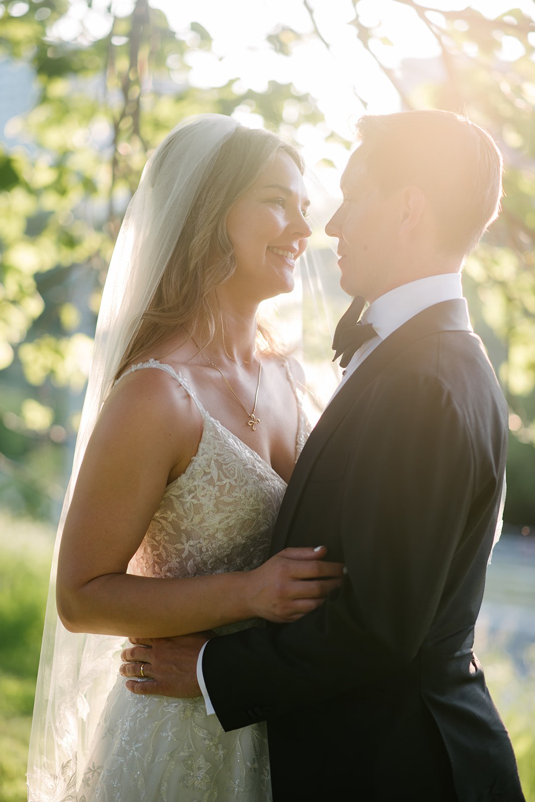 A bride and groom face each other outdoors in sunlight, with trees and leaves behind them. The bride is smiling, wearing a white lace wedding dress and a veil, while the groom is in a black tuxedo with a bow tie. They are holding each other lovingly.