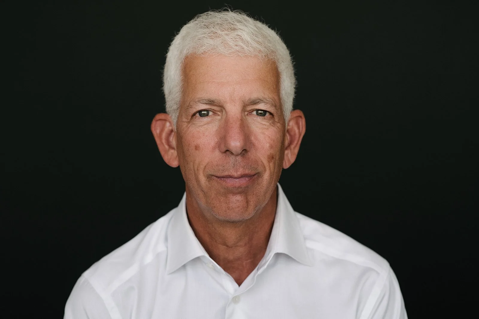 Portrait of a middle-aged man with short white hair, wearing a white collared shirt, against a black background.