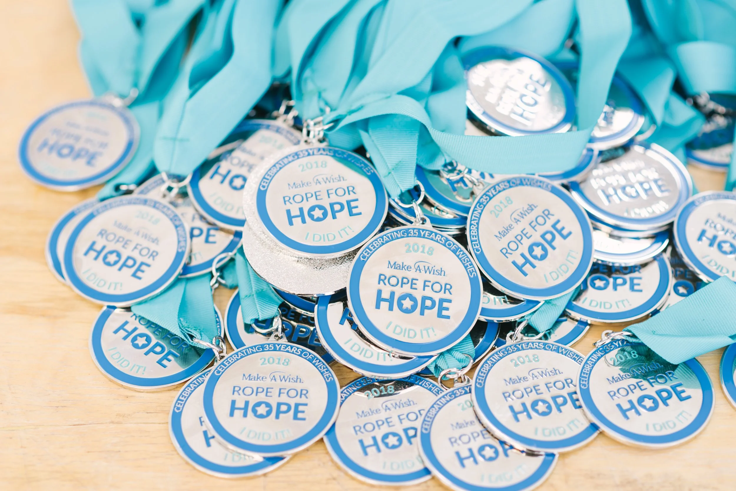 A pile of silver and blue medals with blue ribbons for the 2018 'Rope for Hope' charity event, celebrating 35 years of wishes, displayed on a wooden surface.