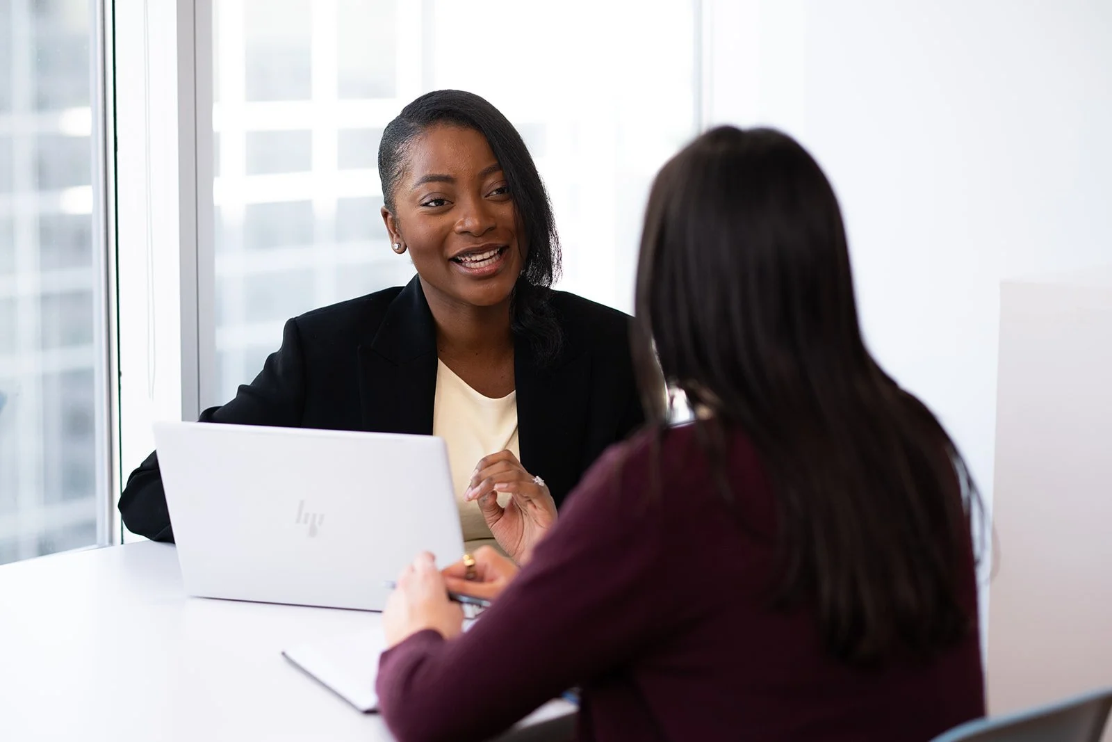 Two women having a conversation in an office, with one woman speaking and the other listening, seated at a white table with laptops.