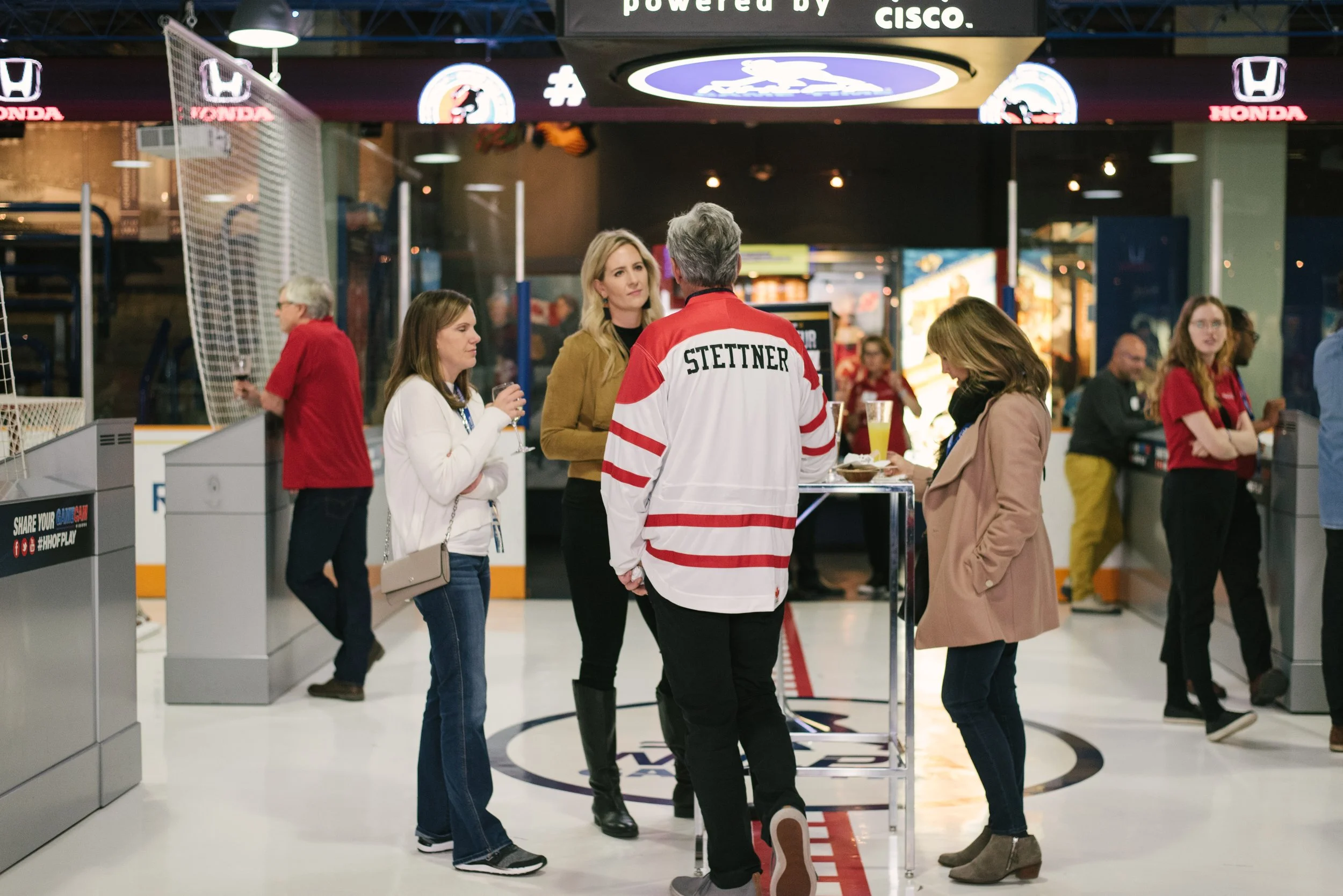 People socializing at Hockey Hall of Fame in Toronto, one person wearing a hockey jersey with 'STETTNER' on the back, others holding drinks, in front of various sports and sponsor signs.