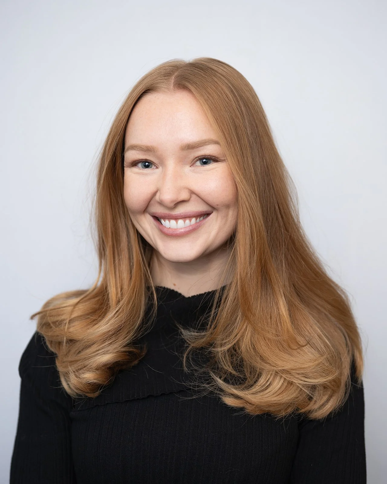 Portrait of a smiling woman with long, wavy red hair wearing a black top against a light gray background.