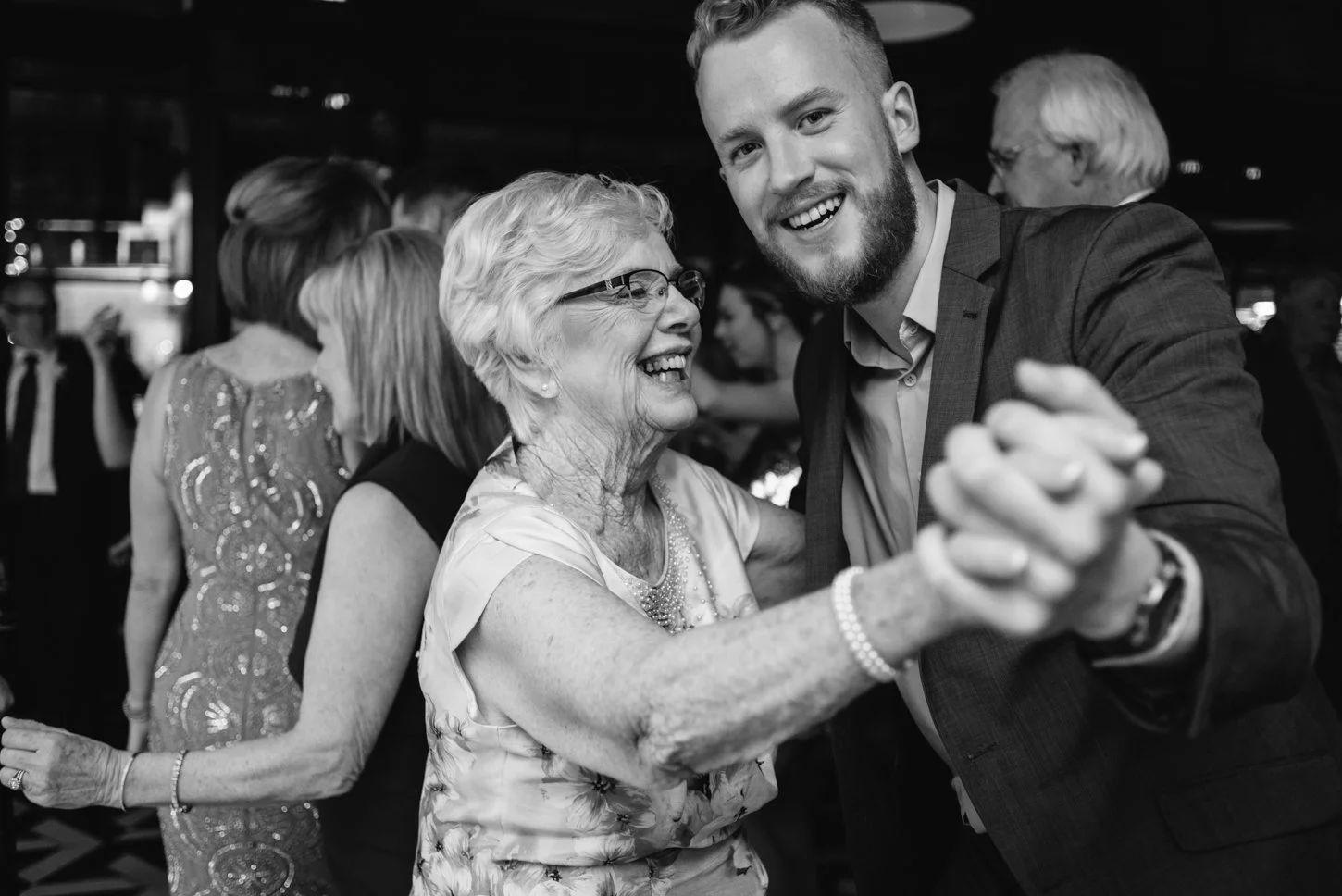 A young man and an elderly woman are dancing and smiling at a social gathering or celebration. The woman is wearing glasses and a floral dress, and the man is dressed in a suit. Other people are in the background, also dancing.
