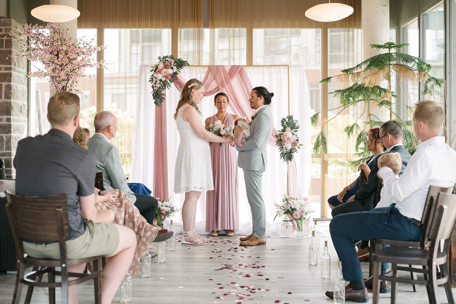 A couple exchanging vows at a wedding ceremony with a pink and white floral backdrop, surrounded by seated guests, indoor setting with large windows, and floral decorations including a cherry blossom branch and potted plants.