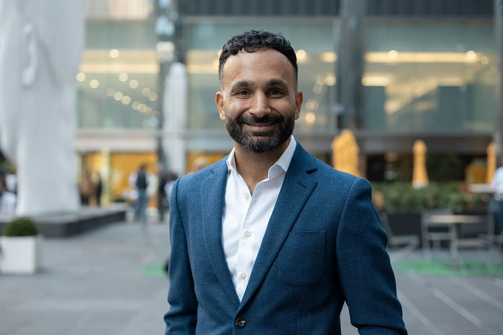 A smiling man in a blue suit and white shirt standing outdoors in an urban setting with buildings and people in the background.