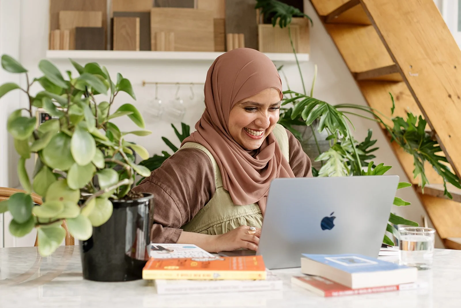A portrait of a woman wearing a pink hijab sitting at a table working on a laptop, surrounded by green plants and books in a cozy, well-lit room.