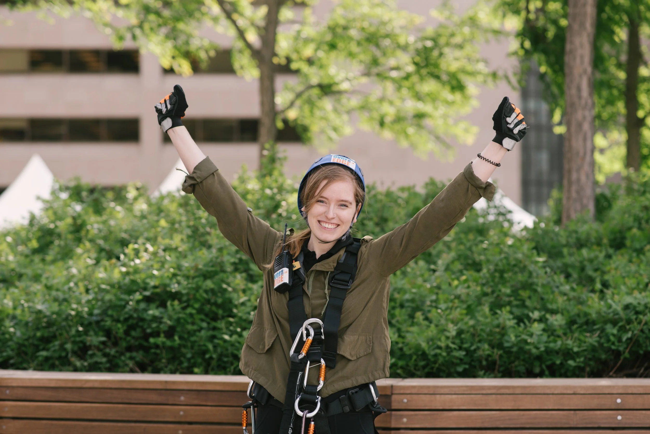 Young woman in outdoor gear with climbing harness, helmet, and gloves smiling with arms raised in park.