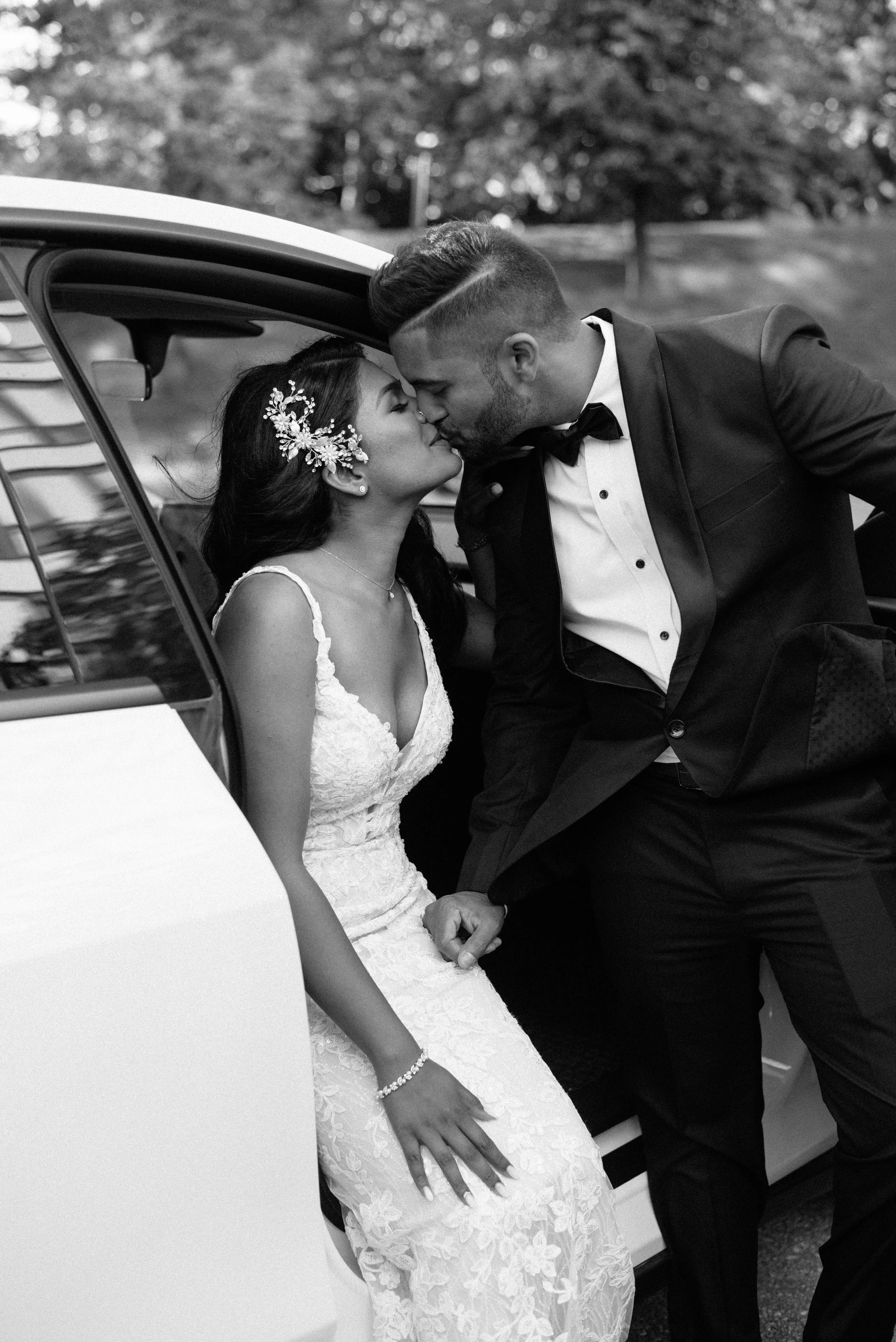 A bride and groom share a kiss next to a car outdoors, black and white photo.