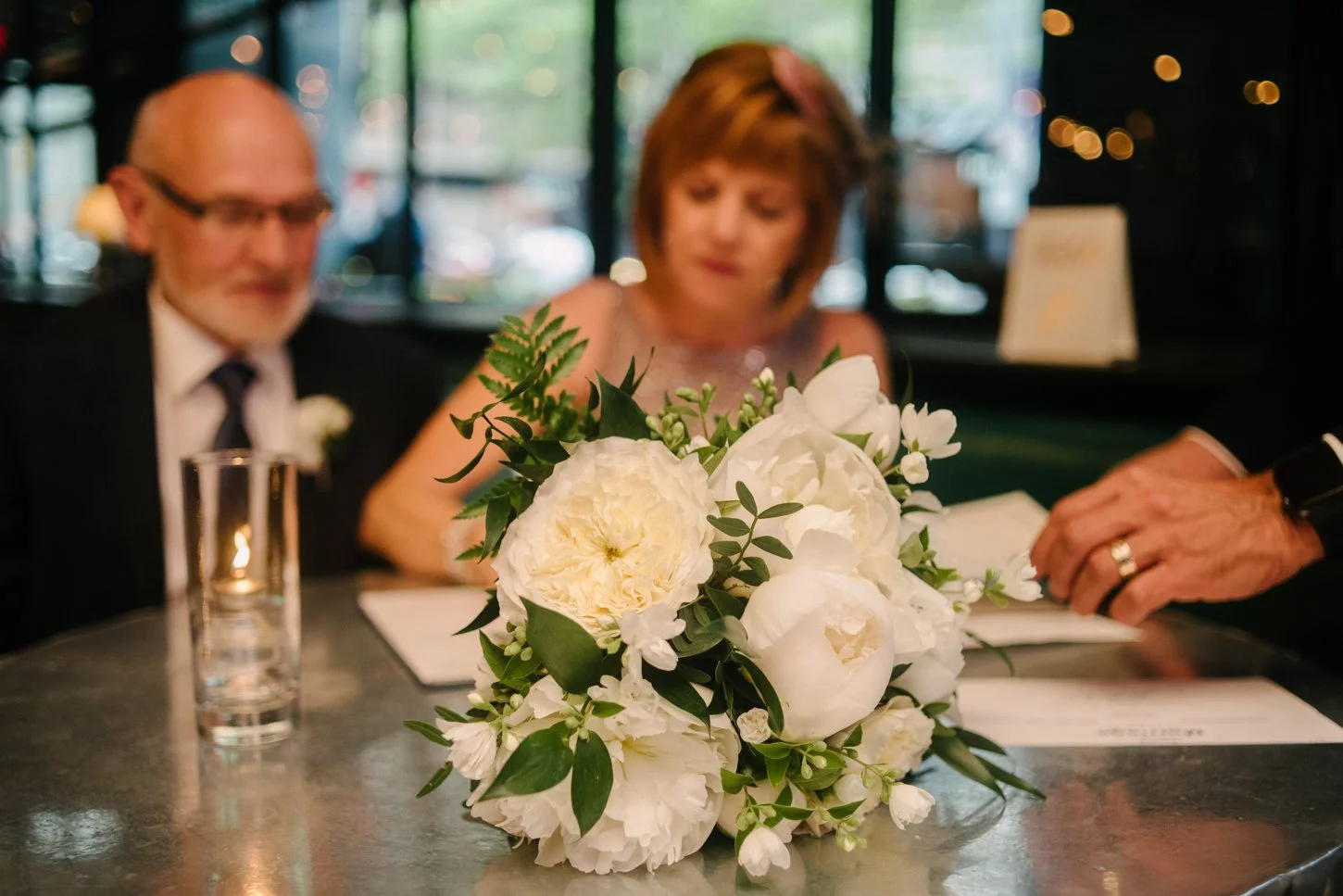 A bouquet of white peony flowers and green leaves placed on a table in a restaurant or cafe, with two blurred people in the background, one man and one woman, and papers or menus on the table.