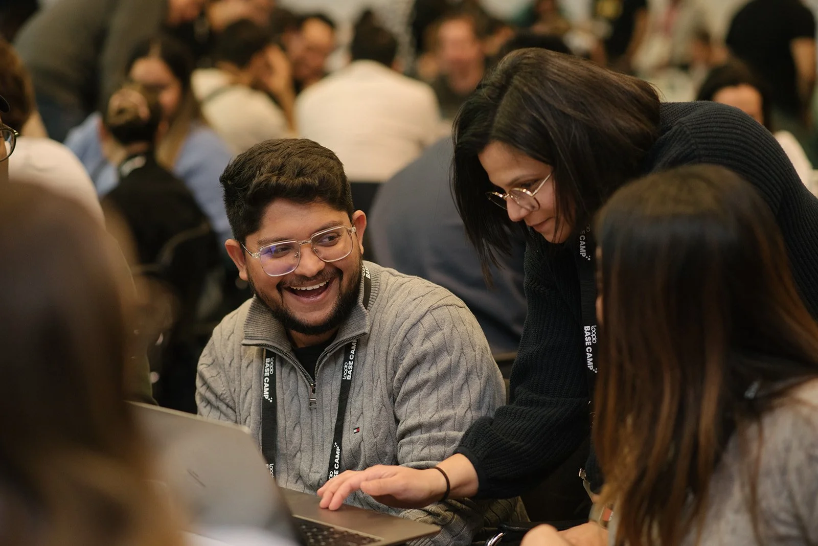A group of people at a conference or workshop, with a young man and woman in the foreground engaging closely and smiling, while others are blurred in the background.
