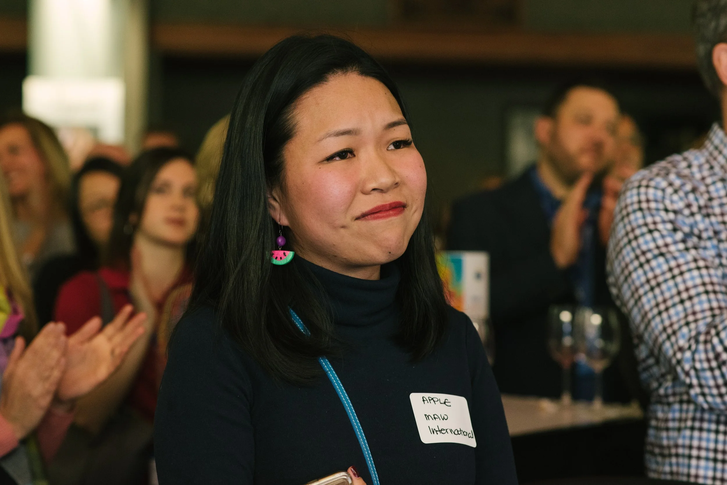 A woman with black hair, wearing a dark top and colorful earrings, attending a formal event with other people in the background.