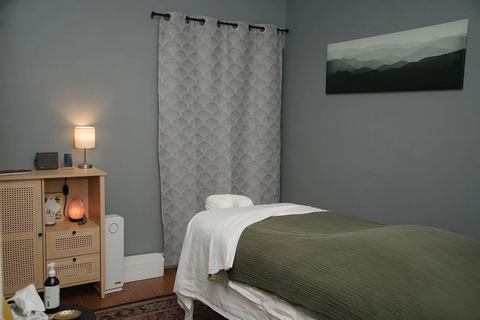 A bedroom with a bed covered in white sheets and a green blanket. There's a gray painted wall, a window with a patterned curtain, a landscape painting, a wooden dresser with a lamp, salt lamp, and small items. A white humidifier and a bottle of hand 