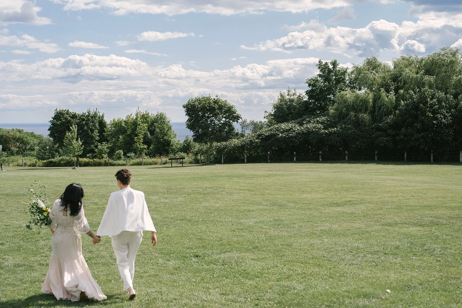 A couple dressed in wedding attire walking hand in hand across a grassy field under a partly cloudy sky, with trees and a distant landscape in the background.