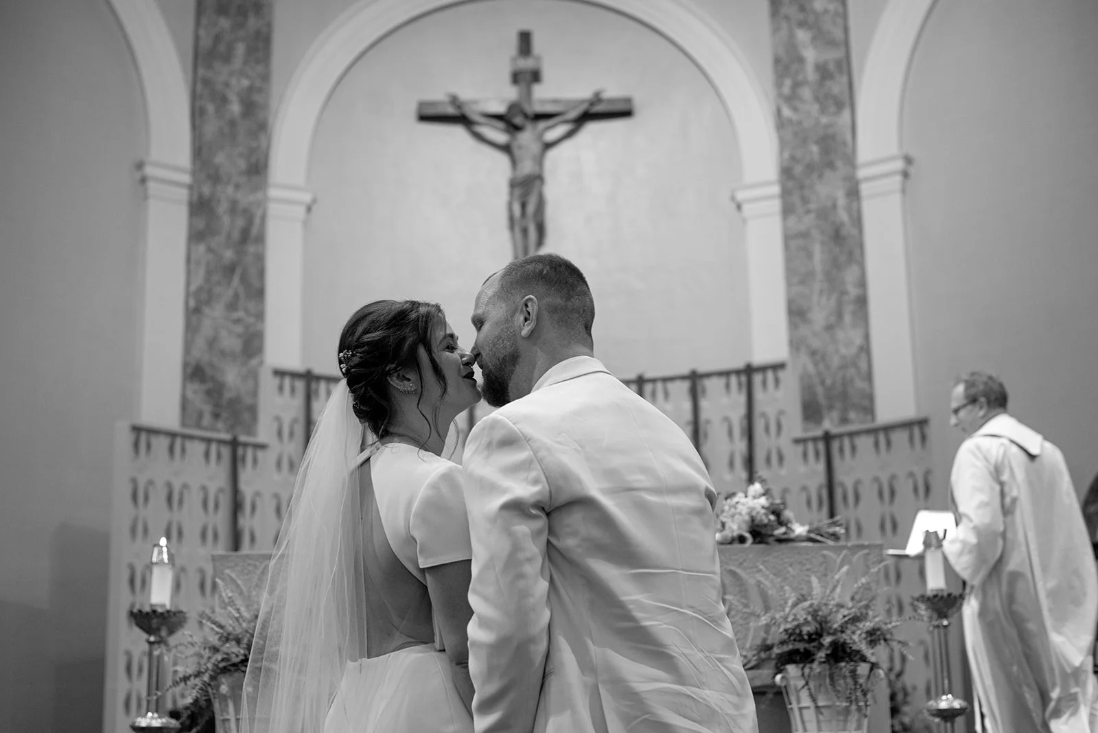 A bride and groom sharing a kiss during their wedding ceremony inside a church, with a crucifix hanging on the wall behind them and a priest in the background.