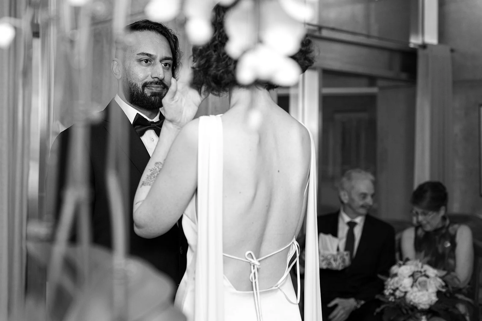 A man and woman are exchanging vows during a wedding ceremony, with the man dressed in a tuxedo and the woman wearing a backless wedding gown, in a decorated indoor setting.