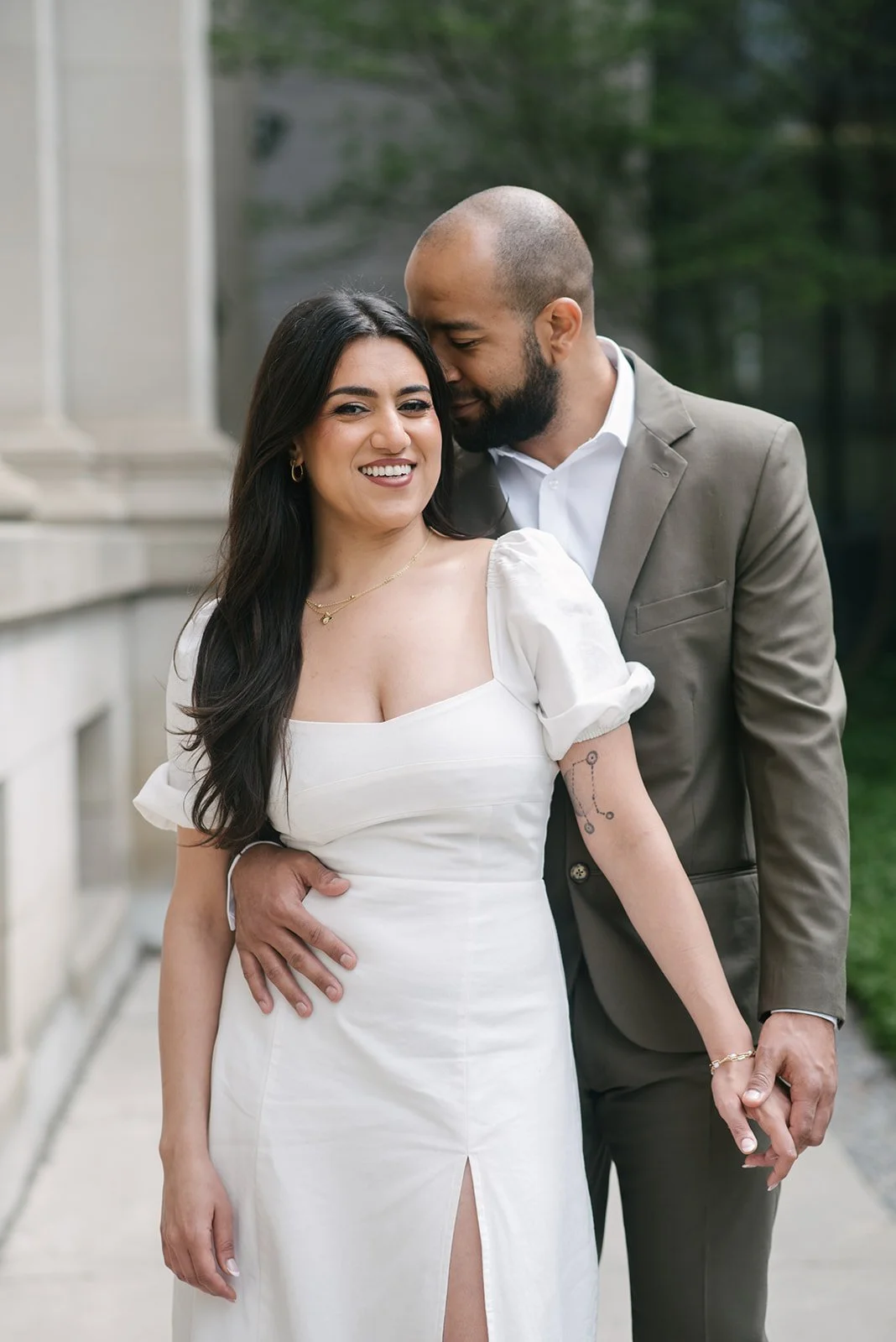 A smiling woman in a white dress with puff sleeves and a slit, holding hands with a man in a gray suit, outside a building with trees in the background, posing for a photo.