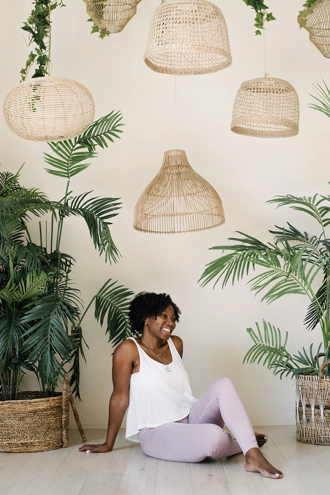 A woman sitting on the floor among large green plants with woven pendant lights hanging above in Mint Room Studios in Toronto.