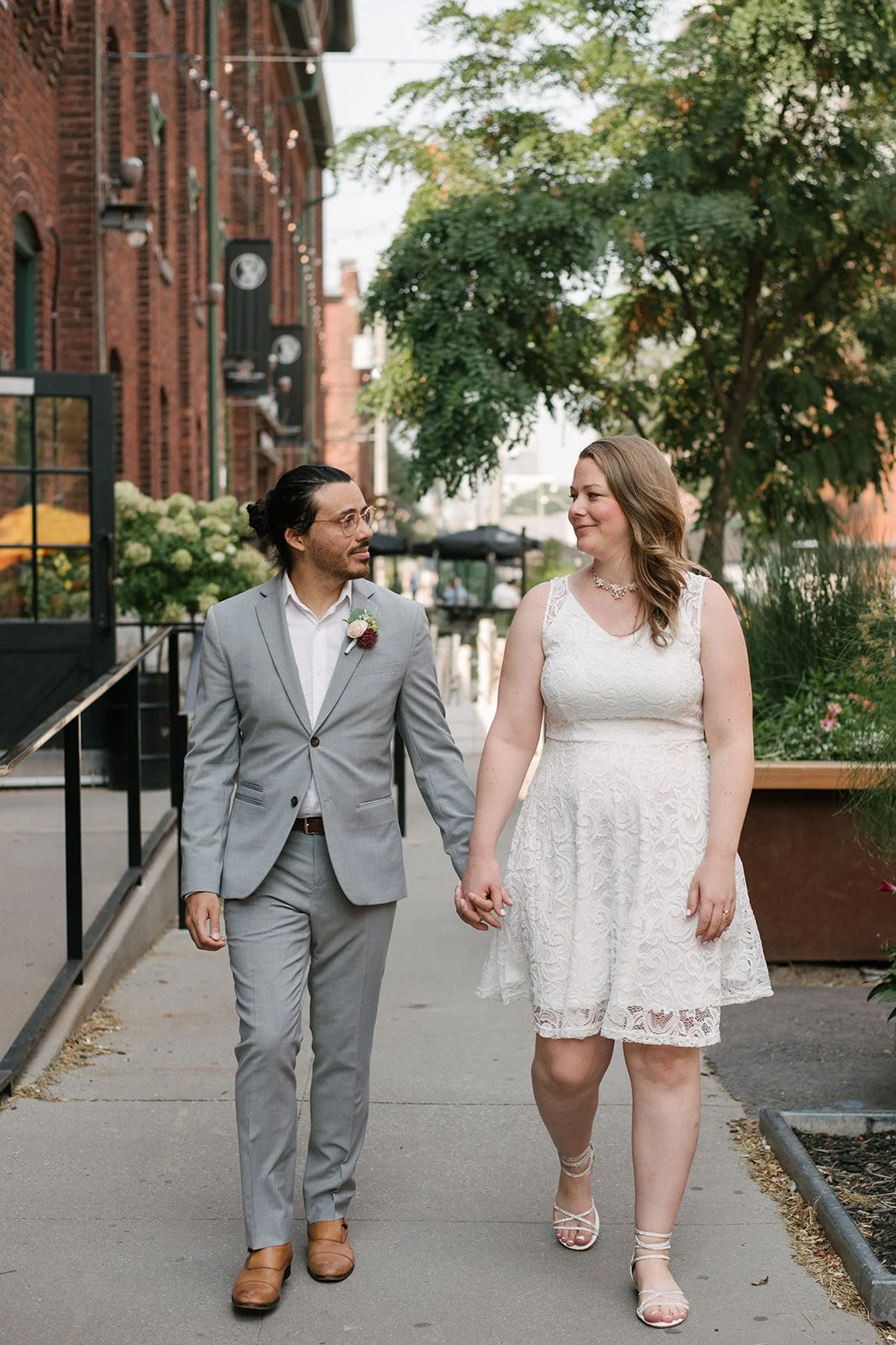 A couple holding hands and walking outdoors in the Distillery District in Toronto, with brick buildings, greenery, and string lights in the background.