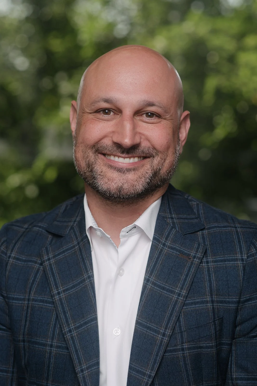 Smiling man with a beard in a checkered blazer and white shirt, standing outdoors with blurred green trees in the background.