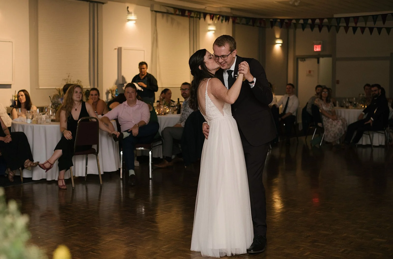 A bride and groom are dancing at their wedding reception, surrounded by seated guests in a decorated banquet hall.