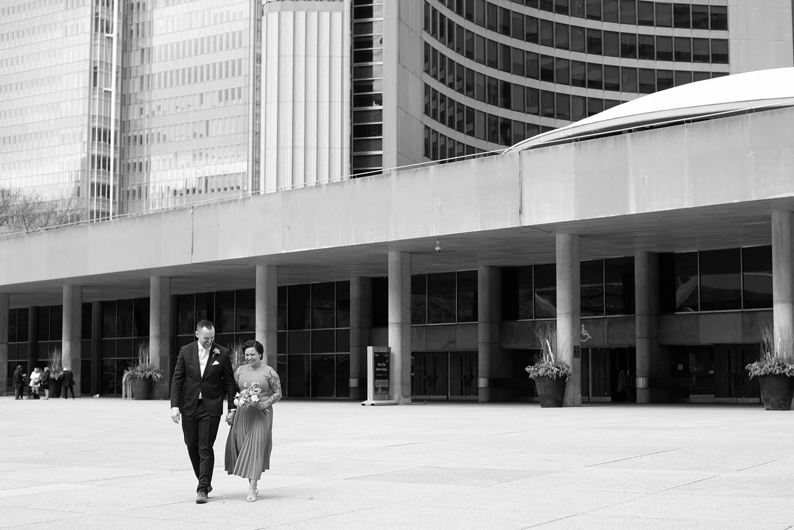 A man and woman walking away from Toronto City Hall, with city hall's building in the background, dressed formally; the woman is a bride and she is holding a small bouquet of flowers.
