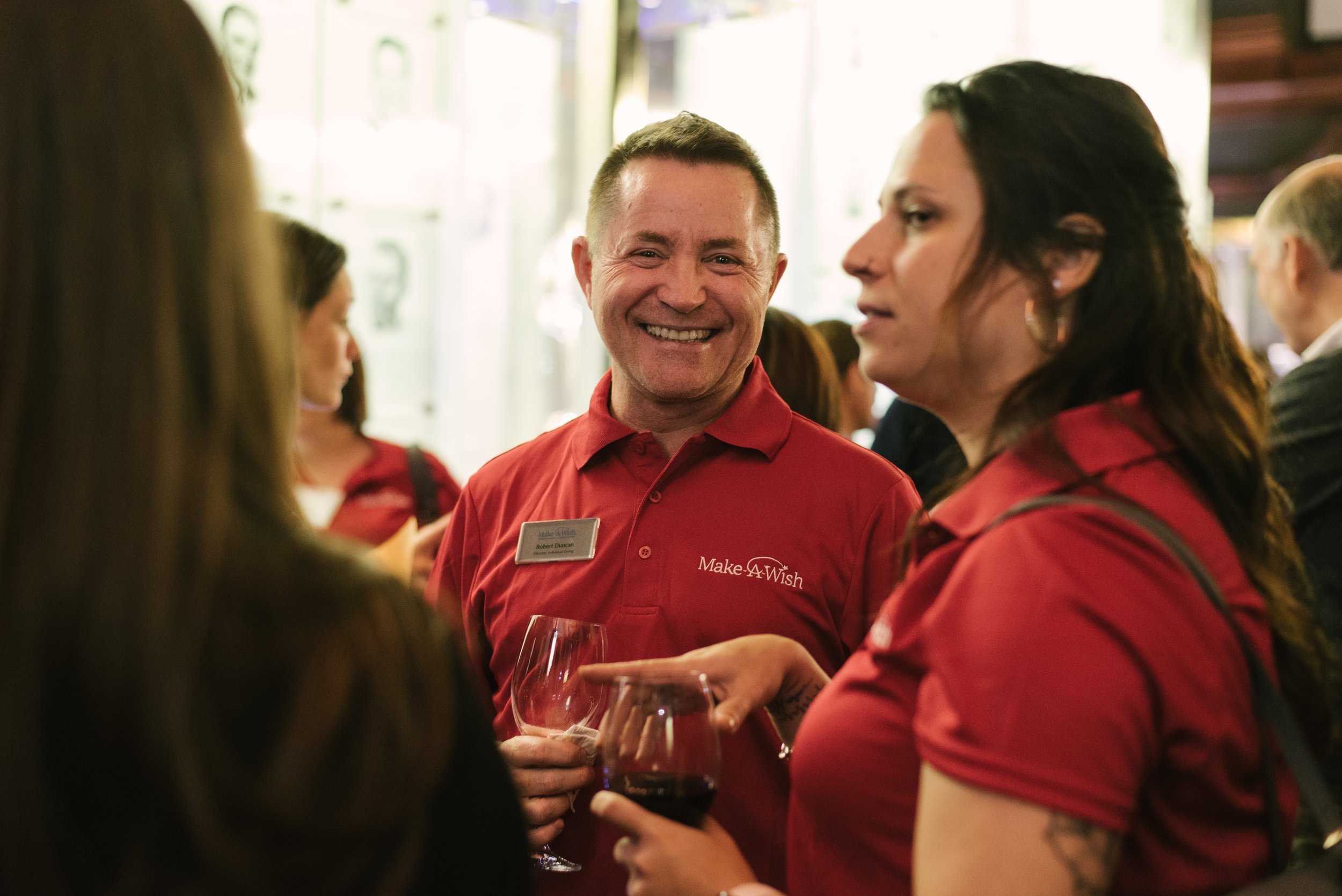 A smiling man in a red 'Make-A-Wish' shirt converses with women at a social gathering, holding a glass of red wine.