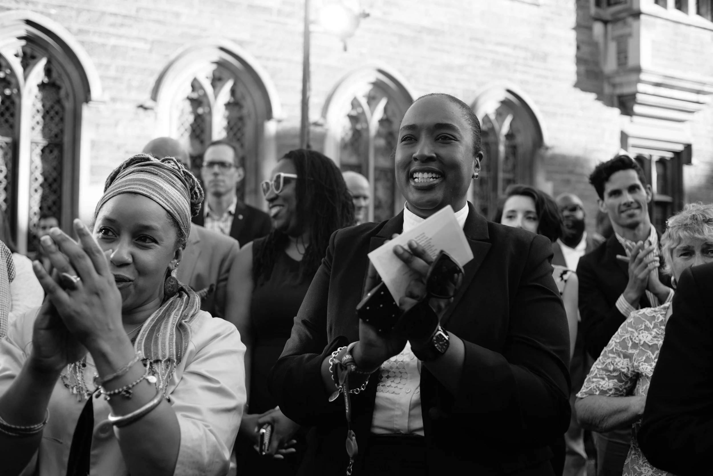 A black and white photo of a group of people attending an outdoor event, with some clapping and smiling, at Hart House. University of Toronto