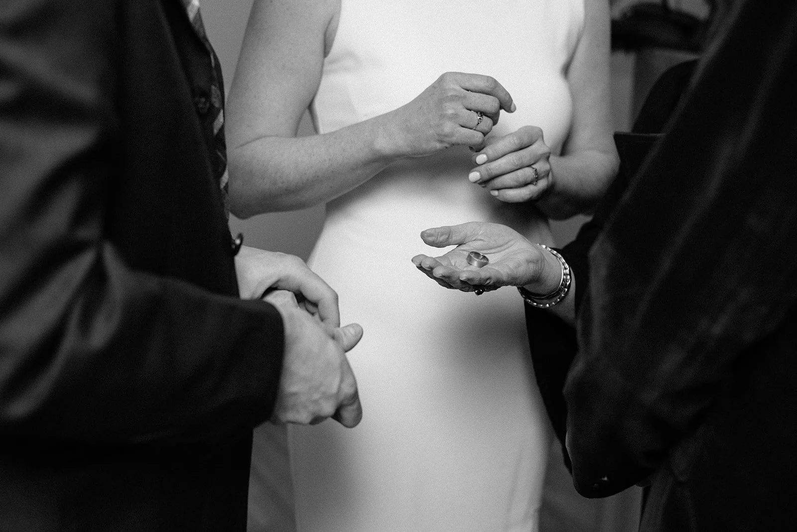 A black-and-white photo of a wedding ceremony, showing a bride and groom exchanging rings with officiant or witness, focusing on hands and rings.