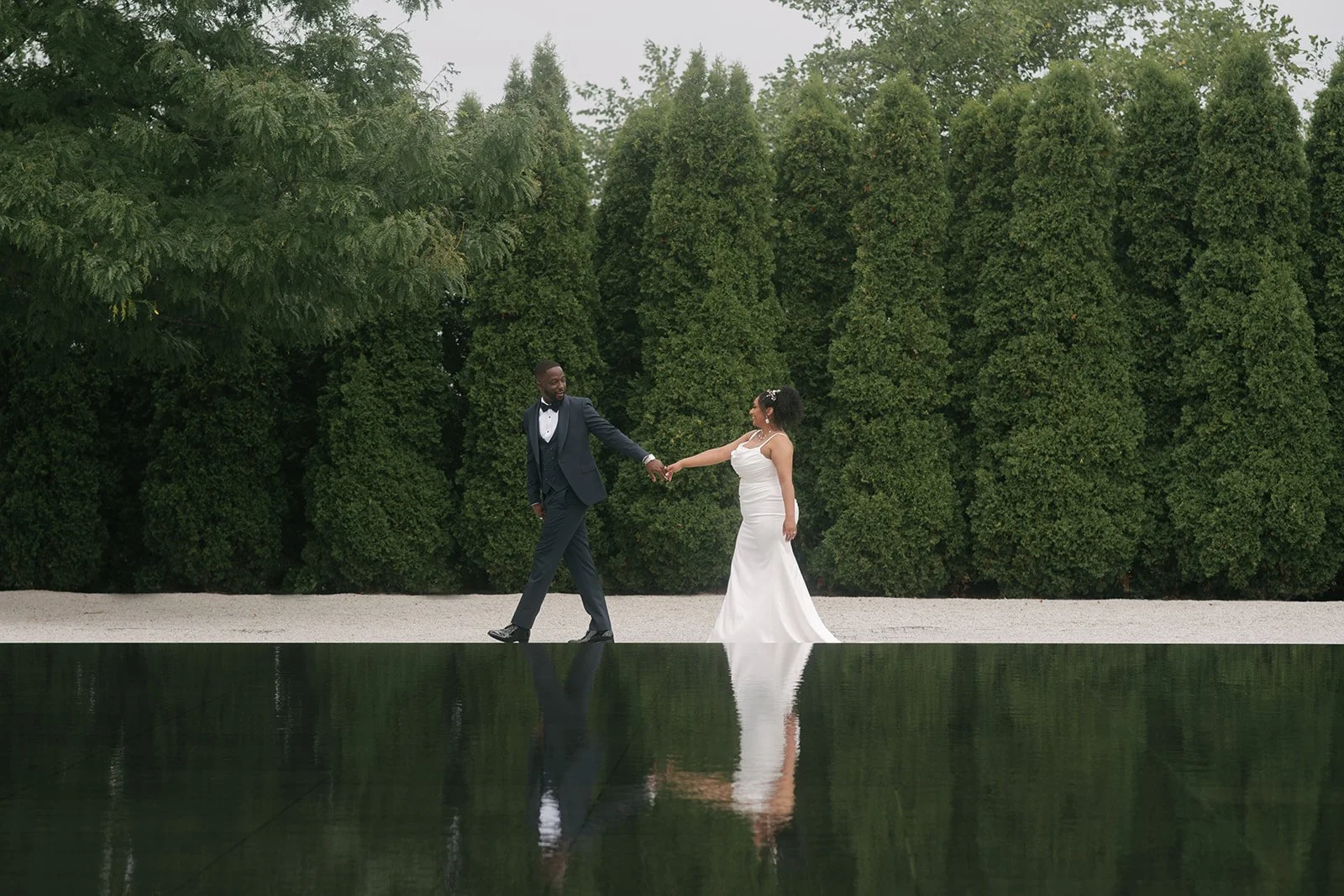 A bride and groom holding hands and dancing outdoors, with a backdrop of tall green trees and a reflective water feature in the foreground.