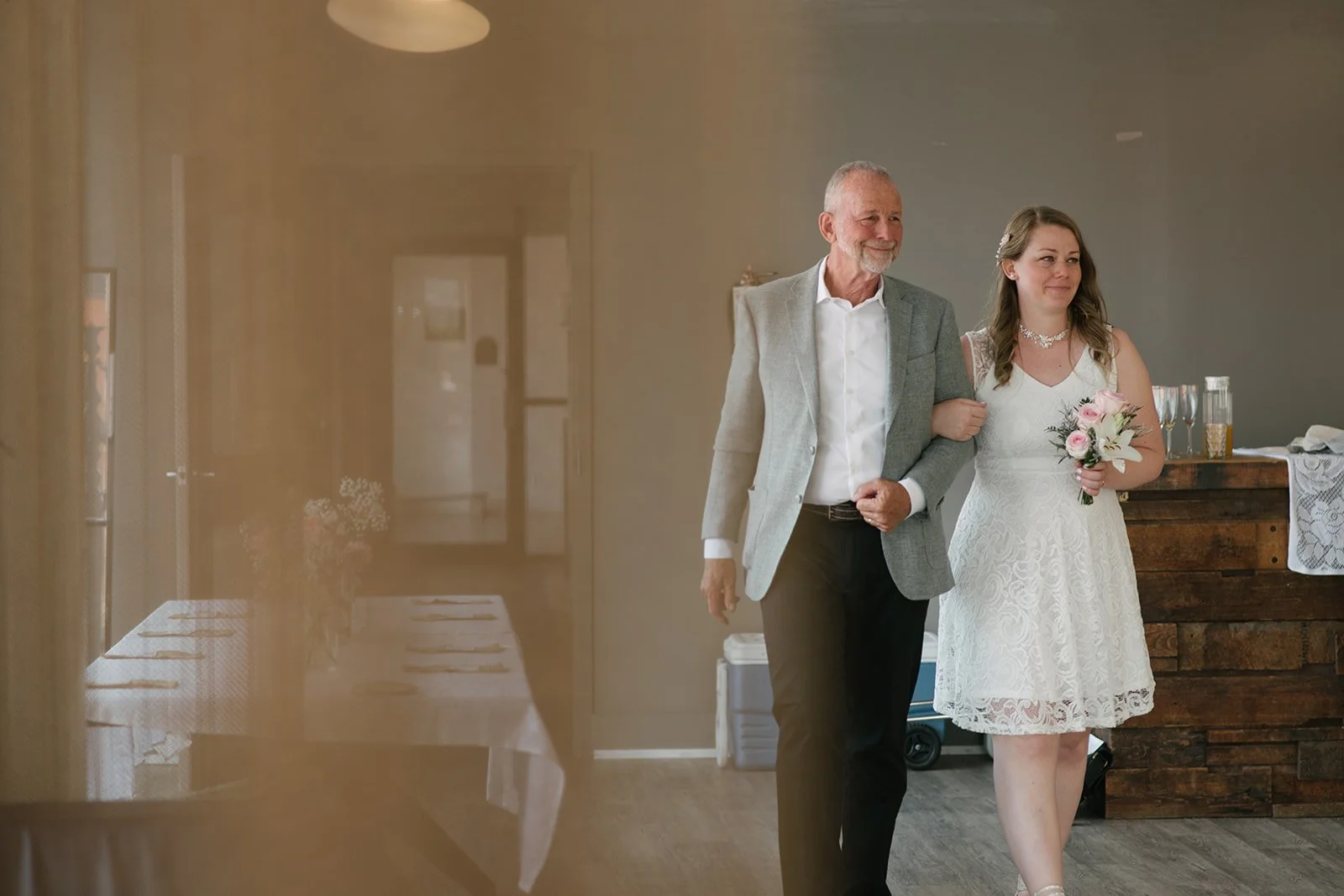 A young woman in a white lace dress holding a bouquet of pink and white flowers walking arm-in-arm with an older man in a gray blazer and white shirt. They are indoors at a wedding reception, with a rustic wooden bar and glasses in the background.