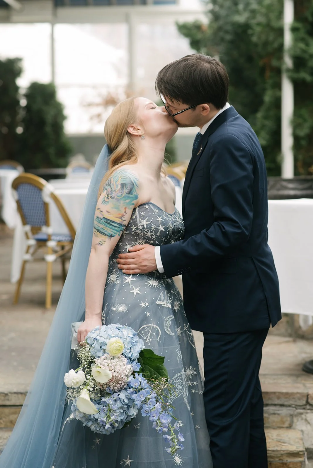 A couple kisses on the bright, indoor terrace at Auberge du Pommier in Toronto. The bride holds a bouquet of white and blue flowers, wearing a starry dress, and the groom in a navy suit.