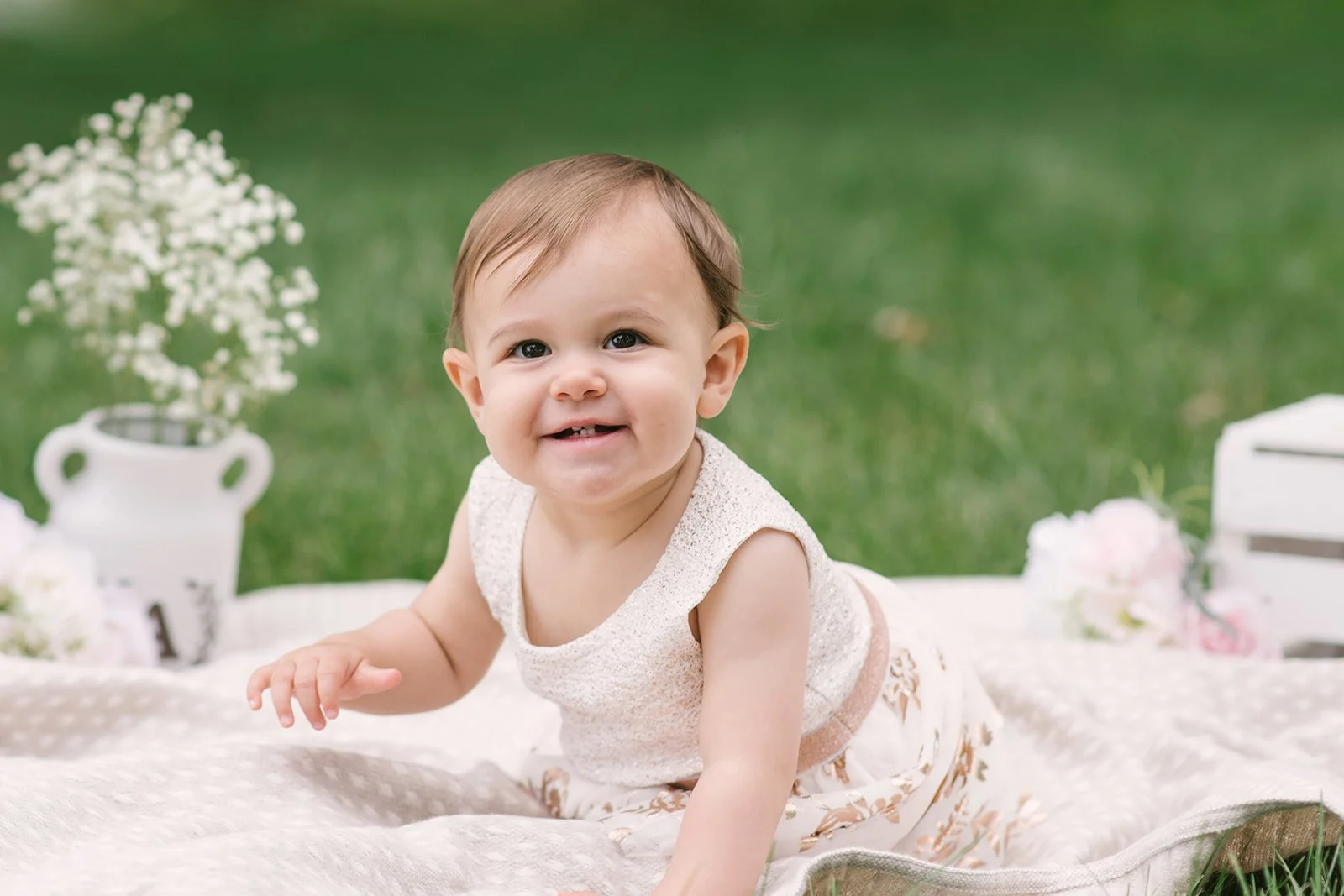 A smiling baby girl crawling on a white blanket outdoors with blurred background of green grass, a white flower arrangement, and decorative items.