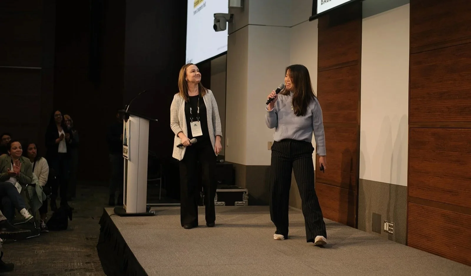 Two women are on stage at a corporate conference, with the woman on the right speaking into a microphone and the woman on the left holding a microphone and looking at her. 