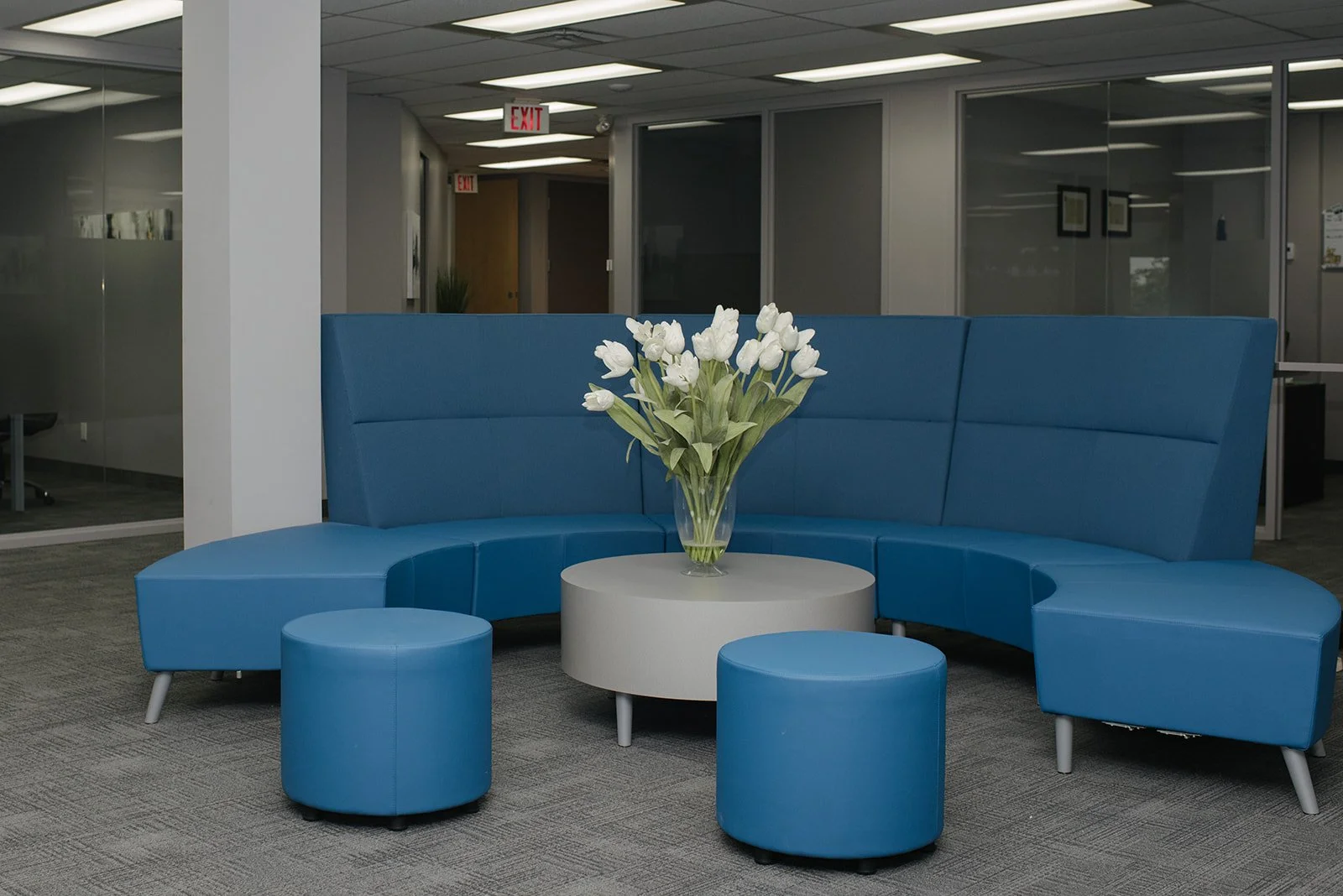 Modern office waiting area with a curved blue sofa, matching blue round ottomans, a white round table with a bouquet of white tulips in a glass vase, glass walls, and closed doors with exit signs.