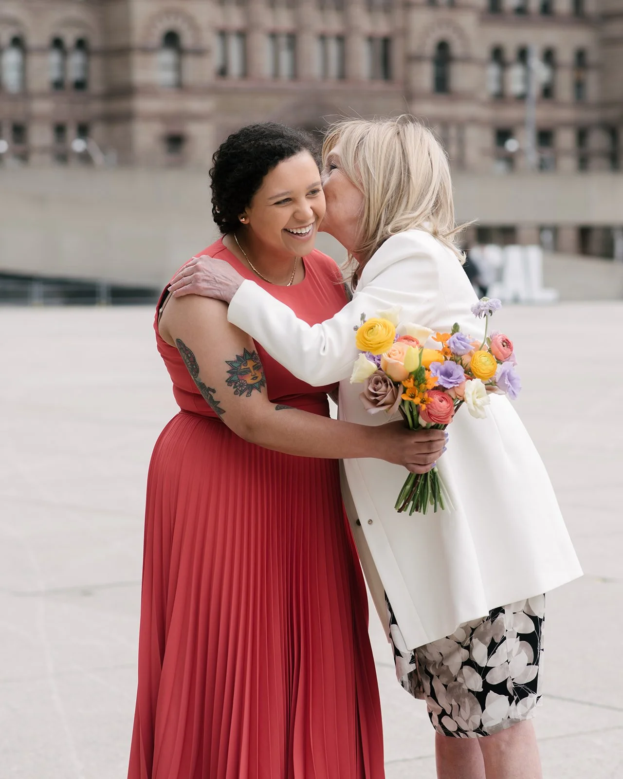 Two women hugging, one a bride who is holding a bouquet of colourful flowers outside of Toronto City Hall.