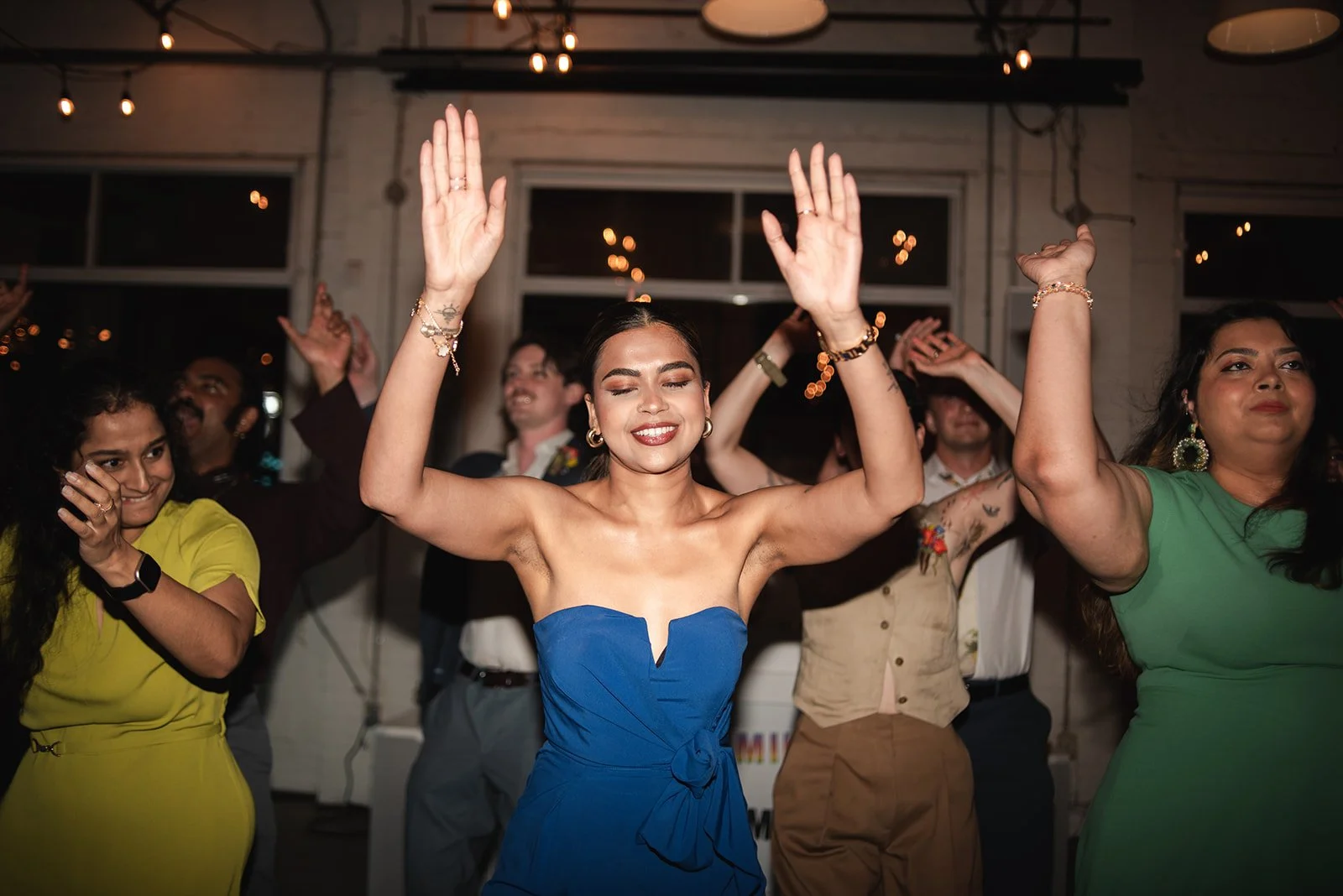 A group of people dancing at a celebration or party, with a woman in a blue strapless dress in the centre raising her hands and smiling.