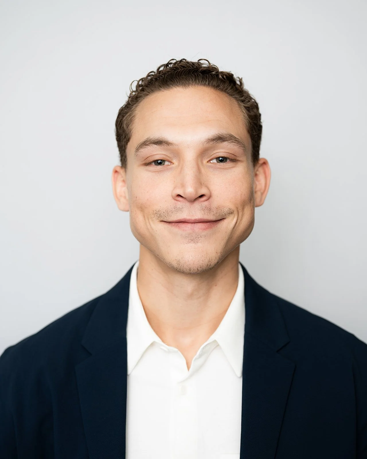 Headshot of a young man wearing a dark suit jacket and white dress shirt, smiling softly against a plain light background.