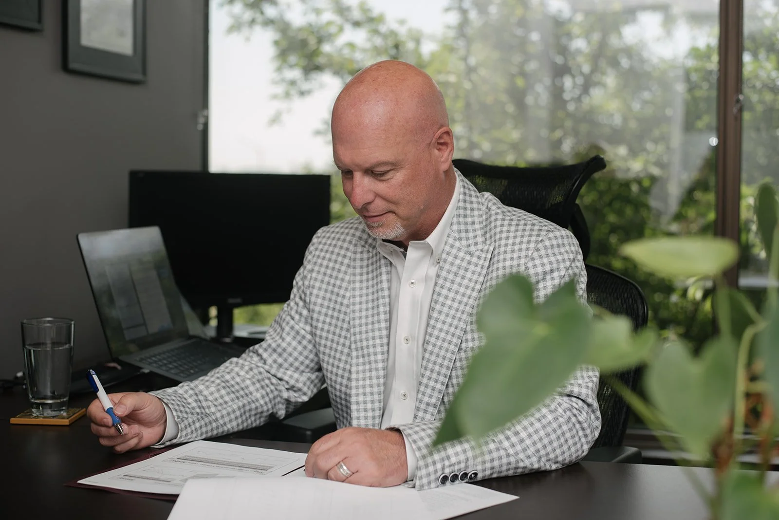 A man in a checkered suit writing on papers at a desk in an office with a large window showing greenery outside.