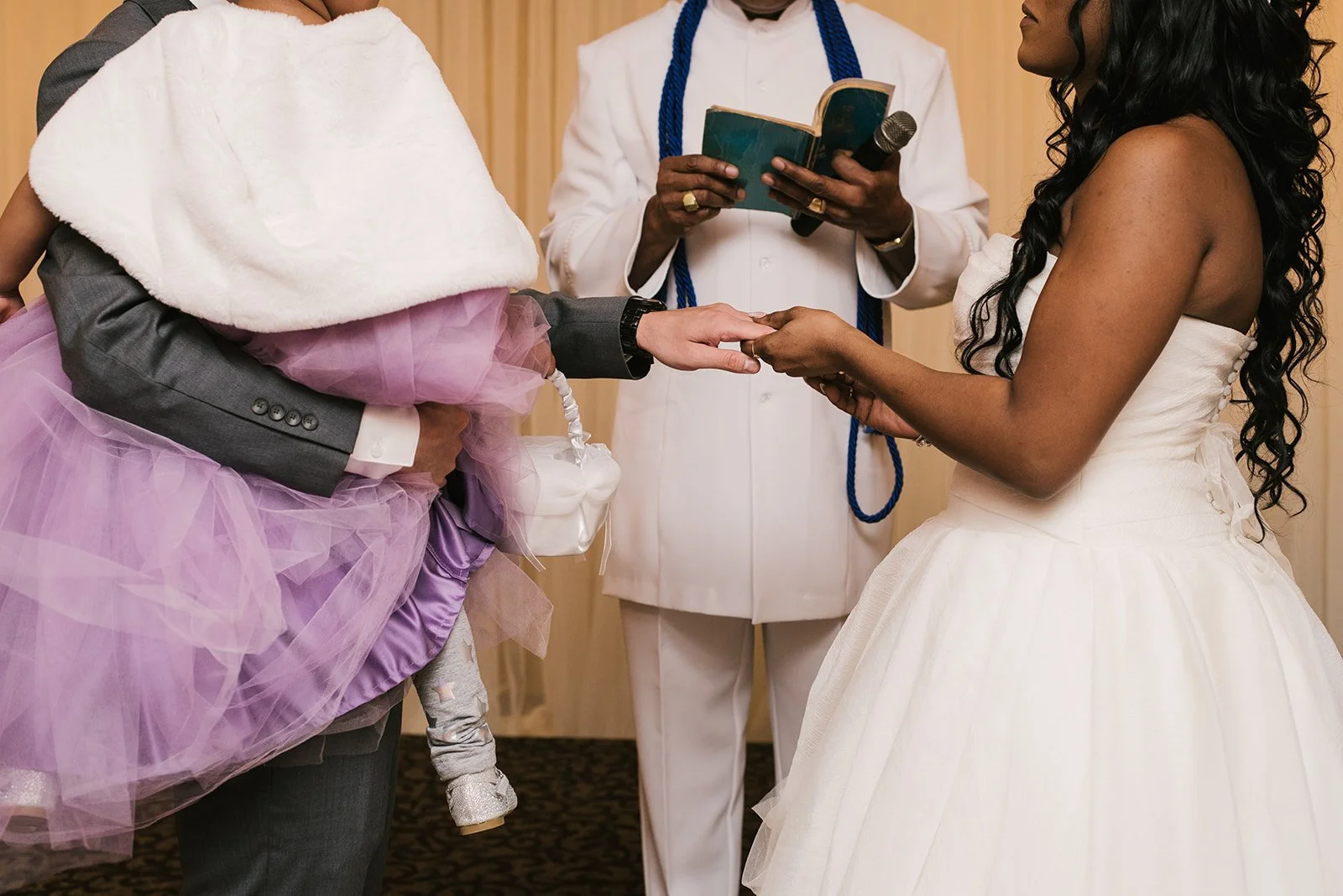 A wedding ceremony with the bride holding hands with the groom, while the groom holds his child in his other arm, and an officiant reading from a book.